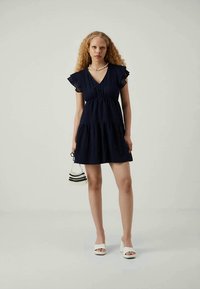 Γυναίκα με σγουρά μαλλιά, φορώντας ένα ναυτικό μπλε φόρεμα με V-λαιμόκοψη, λευκά ανοιχτά παπούτσια με τακούνι, με κολιέ από πέρλες, σκουλαρίκια κρίκους, και κρατώντας μια μικρή τσάντα με χάντρες.