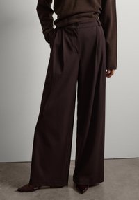 Pantalones de pierna ancha en tejido marrón oscuro, con cintura alta, pliegues, bolsillos laterales y cierre de botón. Textura suave con un corte a medida.