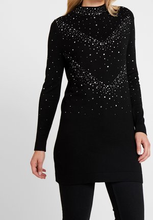 Femme portant une tunique noire à manches longues avec de petites perles blanches éparpillées formant un motif chevron sur le devant et les manches.