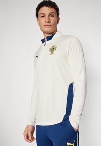 Kreemivärvi pikasleega sportlik pluus, millel on tumesinine krae ja külgpaneelid, ning rinnal kollane rahvuslik embleem ja väike logo.