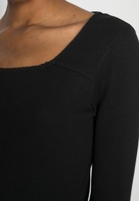 Svart ribbad långärmad topp med ett brett fyrkantigt halsringning och subtilt scalloperade kanter vid kragen, vilket betonar textur och passform.