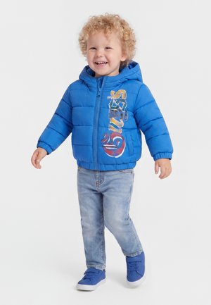 Petit enfant aux cheveux blonds bouclés souriant, portant une doudoune à capuche bleue avec un texte coloré, un jean clair et des baskets bleues, debout sur fond blanc.