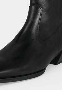 Bottine en cuir noir avec bout pointu et talon bas incliné, montrant des coutures détaillées et une texture lisse.