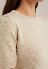 Bežs adīts džemperis ar tekstūru, kurā ir mazi auzu punkti, apaļa kakla izgriezums un īsas piedurknes. Gluda auduma ar ribinātiem piedurkņu galiem.