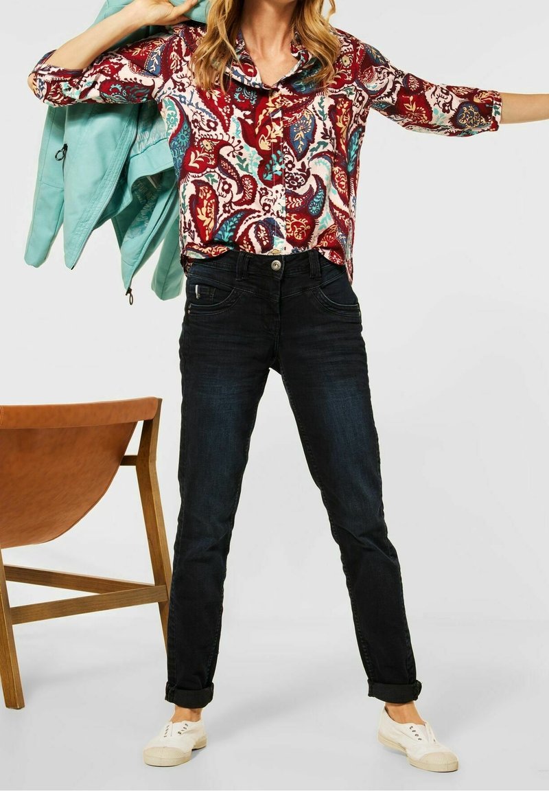 Femme portant un jean noir, des baskets blanches et une chemise colorée à motif cachemire, tenant une veste bleu clair sur son épaule.