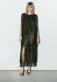 Vrouw draagt een mouwloze zwarte jurk met franjes en een split aan de voorkant, zwarte open-teen hakken en zilveren armbanden, staand tegen een effen muur.