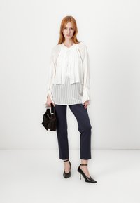 Blusa bianca a righe con fiocco, indossata sopra un top grigio a righe. I pantaloni blu navy, i tacchi alti neri e una piccola borsa scura completano l'outfit.