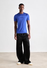 Sininen lyhythihainen T-paita, jossa on pyöreä kaula-aukko ja Calvin Klein -logo, yhdistettynä leveisiin mustiin housuihin ja vaaleisiin slip-on -kenkiin.