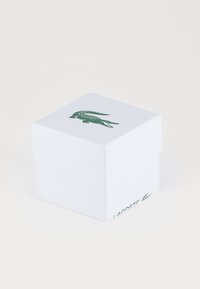 Baltas kvadratinis laukelis su tekstūruota paviršiumi, puoštas žaliu krokodilo logotipu ir žodžiu "LACOSTE" žalia spalva apačioje.