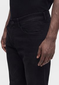 Person trägt schwarze Jeans und ein schwarzes Shirt, steht mit einem Arm entspannt an der Seite vor einem schlichten, hellen Hintergrund.