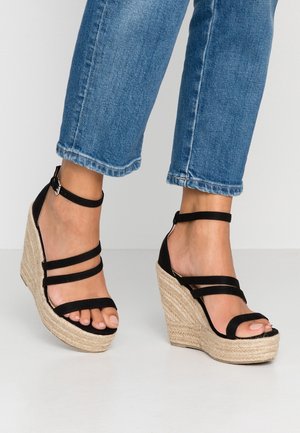 Pieds portant un jean bleu et des sandales compensées noires à lanières avec des talons plateforme en jute tressé sur un sol blanc.
