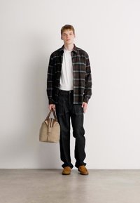 Junger Mann in einem schwarzen karierter Hemd, weißem T-Shirt, schwarzen Hosen und braunen Schuhen, der eine beige Reisetasche gegen eine schlichte weiße Wand hält.