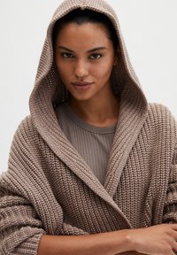 Lange gebreide cardigan in zacht taupe, met een grote capuchon, open voorkant en geribbelde structuur. Gestyled over een aansluitende grijze top.