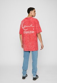 Red, mineral-washed oversized t-shirt with white "Kani" logo and "karl kani-jeans" text on the back. Light blue jeans and black sneakers worn.