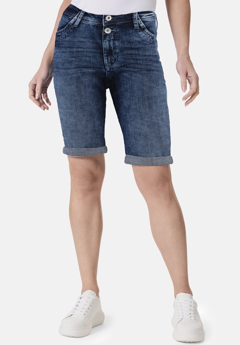 Denim-Shorts mit einer dunkelblauen Waschung, hochgekrempelten Säumen, zwei vorderen Knöpfen und traditionellem Fünf-Taschen-Design. Kombiniert mit weißen Sneakers.