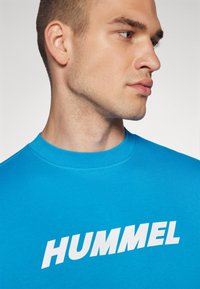 Jasnoniebieska koszulka z okrągłym dekoltem, wykonana z bawełny. Posiada duży, biały napis "HUMMEL" w pogrubionej, ukośnej czcionce na przodzie. Gładka faktura.