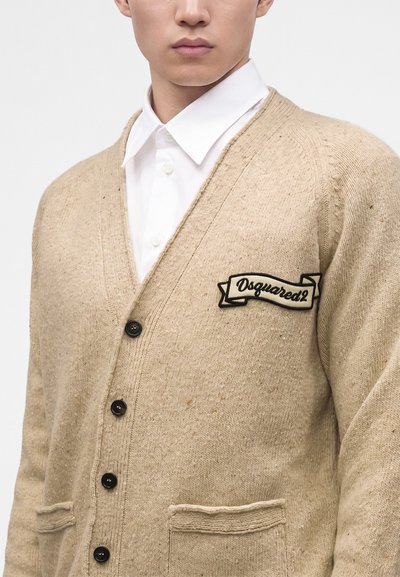 Personne portant un cardigan tricoté beige avec des boutons noirs, des poches avant et un patch "Dsquared2" sur une chemise blanche à col.