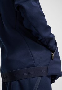 Marinblå sportjacka med mjuk textur, en ficka med dragkedja och en elastisk midjerem med logotyp för en figurnära design.