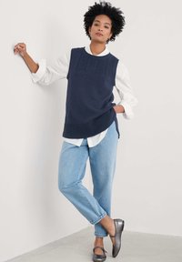 Suéter sin mangas de punto en color marino con parte superior texturizada, combinado con una camisa blanca de botones y jeans ajustados azul claro. Zapatos planos grises.