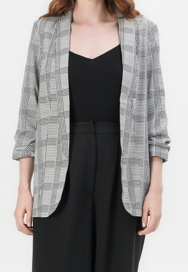 Blazer à carreaux gris avec manches retroussées et ouverture devant, en tissu texturé avec un intérieur noir contrastant. Porté sur un haut noir.