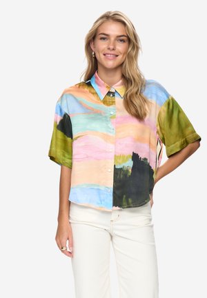 Femme souriante aux longs cheveux blonds, portant une chemise multicolore à manches courtes boutonnée et un pantalon blanc, debout avec une main sur la hanche.