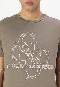 Brown cotton t-shirt featuring a white graphic design with interlocking letters and the text "GUESS?, INC. CLASSIC DESIGN" below.