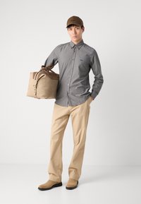 Bolsa de viaje beige con acentos en marrón, camisa gris de botones, pantalones caqui y gorra marrón. Texturas suaves y elementos de diseño casual.