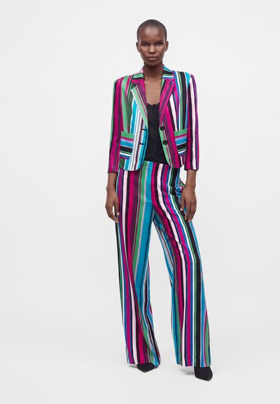 Personne grande portant un blazer à rayures verticales colorées et un pantalon large assorti, avec un haut noir et des chaussures noires, debout sur fond blanc.
