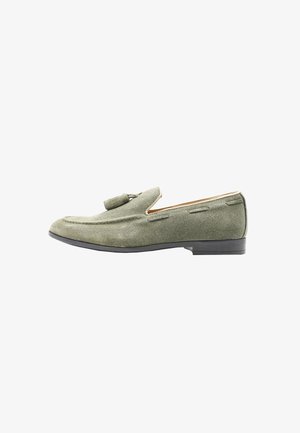 Mocassino da uomo in suede verde oliva con nappe, cuciture dettagliate e tacco basso nero, mostrato di profilo su sfondo bianco.