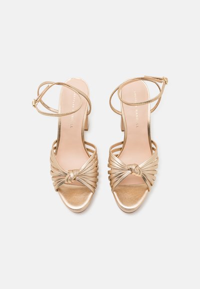 Sandalias de tacón alto doradas con un diseño trenzado, un acento anudado en la parte delantera y correas delgadas en el tobillo. Textura metálica suave.