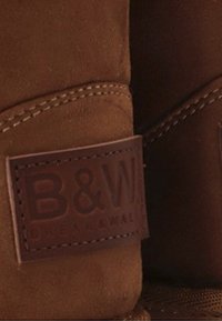 Botas de ante marrón con costuras visibles y una etiqueta rectangular de cuero grabada con "B&W" y un texto más pequeño debajo.