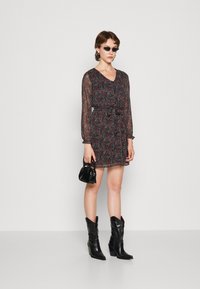 Vestido preto com padrão floral, mangas longas transparentes, cintura ajustada e punhos franzidos. Usado com botas de cowboy pretas e uma pequena bolsa.