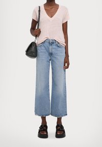 Osoba w jasnoróżowej bluzce z dekoltem w serek, krótkich rękawach, szerokich, wyciętych niebieskich dżinsach, czarnych platformach i nosząca czarną torbę na ramię.