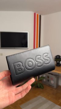 Črn usnjeni denarnica z vtisnjenim logotipom "BOSS", pravokotne oblike, gladke teksture in čistimi šivi. Držana v roki proti ozadju.