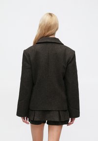 Barna texturált blézer széles gallérral, egyenes szabású és hosszú ujjú, egy hozzáillő rövid szettre viselve. Egyszerű hátkialakítás.