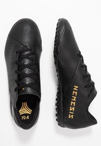 Botas de fútbol negras con una superficie texturizada, que presentan líneas tonales, cordones negros y un logo dorado de "NEMESIS". Diseñadas para el rendimiento y la tracción.