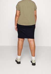 Persona de pie de espaldas, vistiendo una blusa verde oliva, una falda negra hasta la rodilla, calcetines blancos y zapatos beige con suelas negras.