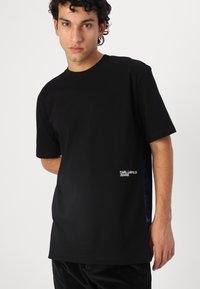 Black cotton t-shirt featuring short sleeves, a crew neck, and a logo detail in white on the lower left side. Smooth texture, relaxed fit.