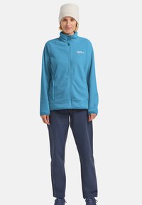 Giacca in fleece blu chiaro con colletto alto, due tasche laterali e zip, abbinata a pantaloni blu navy e a un berretto color crema.