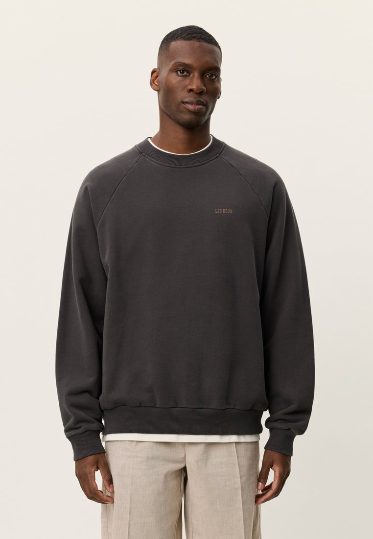 Άνδρας που φοράει σκούρο γκρι φούτερ με λαιμόκοψη και το κείμενο "LES DEUX", μπεζ παντελόνι, στέκεται μπροστά σε ένα απλό φωτεινό φόντο.