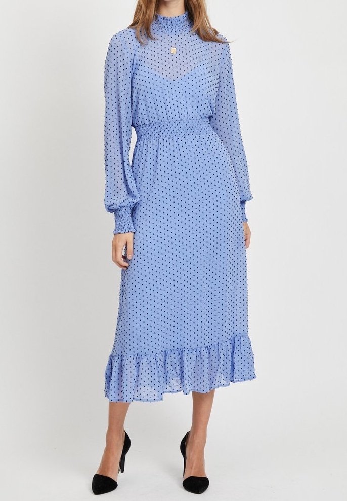Femme portant une robe midi bleu clair à manches longues, à pois noirs, avec une taille et des poignets froncés, un ourlet volanté, et des talons noirs pointus.