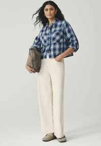 Vrouw met lang krullend haar, gekleed in een blauw geruite blouse, witte wijde broek, beige schoenen en een grote bruine handtas.