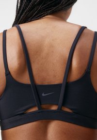Svart sports-bh med tynne stropper, utskjæringsdetalj bak, og liten grå Nike-logo. Laget av glatt, stretchy stoff for komfort.