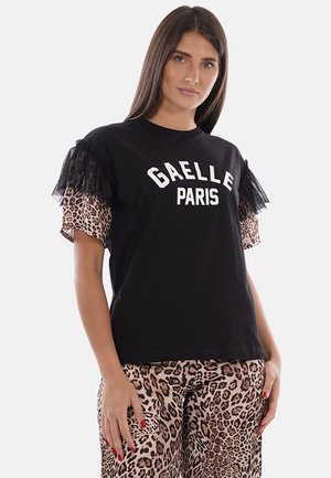 Donna che indossa una T-shirt nera "GAELLE PARIS" con maniche leopardate e pantaloni leopardati abbinati, in piedi su sfondo bianco.