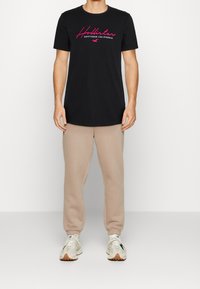 Svart bomullströja med rosa texten "Hollister Southern California", kombinerad med beige joggers och vita sneakers. Slät textur, avslappnad passform.