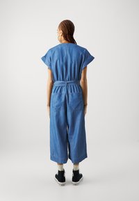 Combinaison en denim à manches courtes, taille ceinturée, design à jambes larges, et deux poches arrière. Portée avec des baskets montantes noires.