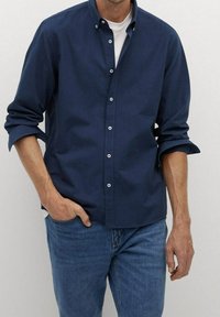 Granatowa koszula na guziki z długimi rękawami, wykonana z lekkiego materiału; ma klasyczny kołnierzyk i białe guziki, noszona na białej koszulce.