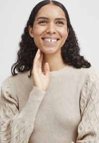 Beige Strickpullover mit geripptem Körper und strukturierten Kabelstrickärmeln. Das Design umfasst einen runden Halsausschnitt und eng anliegende Bündchen.