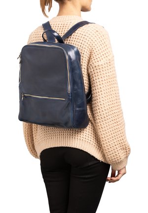 Zaino in pelle blu navy con forma rettangolare, tasca frontale con zip e spallacci imbottiti, indossato sopra un maglione in maglia color crema.