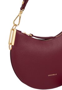 Borsa a tracolla in pelle bordeaux con forma curva, dettagli in metallo dorato e chiusura con zip. Presenta un logo in rilievo.